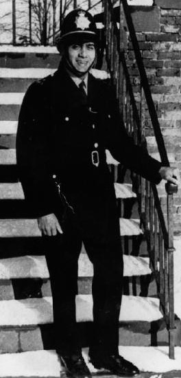Birmingham's first Black Police officer, Ralph Ramadhar stands at the bottom of a flight of stairs. He is dressed in 1960s police uniform and smiling at the camera.